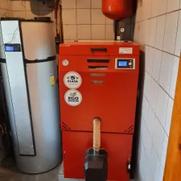 Czerwony, automatyczny kocioł na paliwo stałe, oznaczony naklejkami '5 Klasa' i 'Eco Design', umieszczony w kotłowni z białymi płytkami, obok srebrnego zasobnika na wodę.