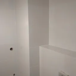 Świeżo ułożone, białe, błyszczące płytki ceramiczne na ścianach w narożniku łazienki, widoczne otwory pod instalacje i wystający fragment ściany tworzący półkę, gniazdko elektryczne w dolnym rogu.