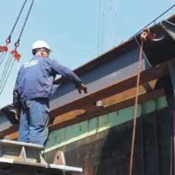 Pracownik w kasku i roboczym ubraniu ZUSO, stojący na platformie, nadzoruje montaż stalowej belki konstrukcyjnej przy użyciu żurawia na tle błękitnego nieba.