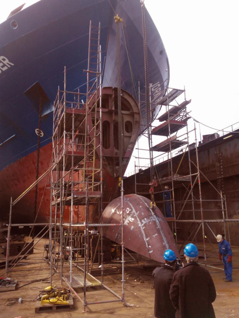 Prace spawalnicze przy kadłubie statku w doku, widoczne rusztowania, spawarki oraz fragment nowej części kadłuba podwieszonej na łańcuchach.