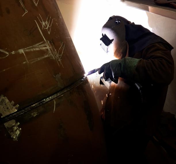 Spawacz w masce spawalniczej łączy dwa elementy metalowe o zakrzywionej powierzchni, widoczne iskry i ślady po kredzie na metalu.