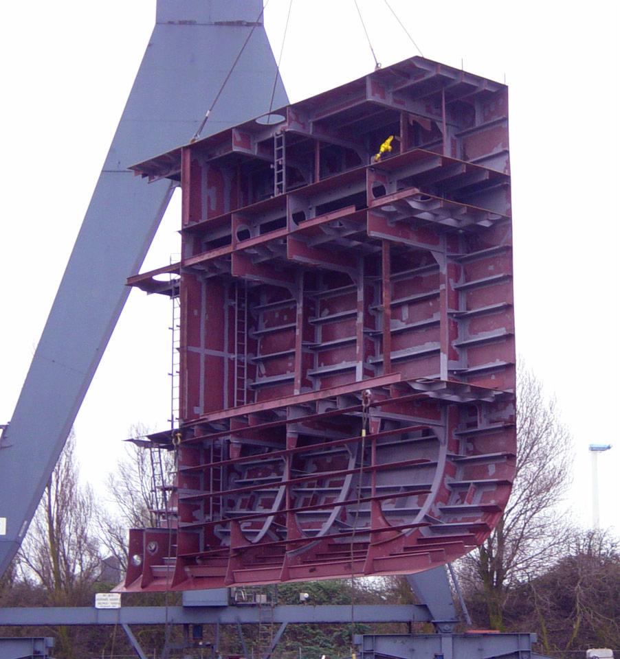 Fragment kadłuba statku w trakcie budowy, podnoszony przez dźwig, widoczna konstrukcja stalowa z licznymi wzmocnieniami i drabinką, pracownik w żółtym kombinezonie na górnym poziomie.