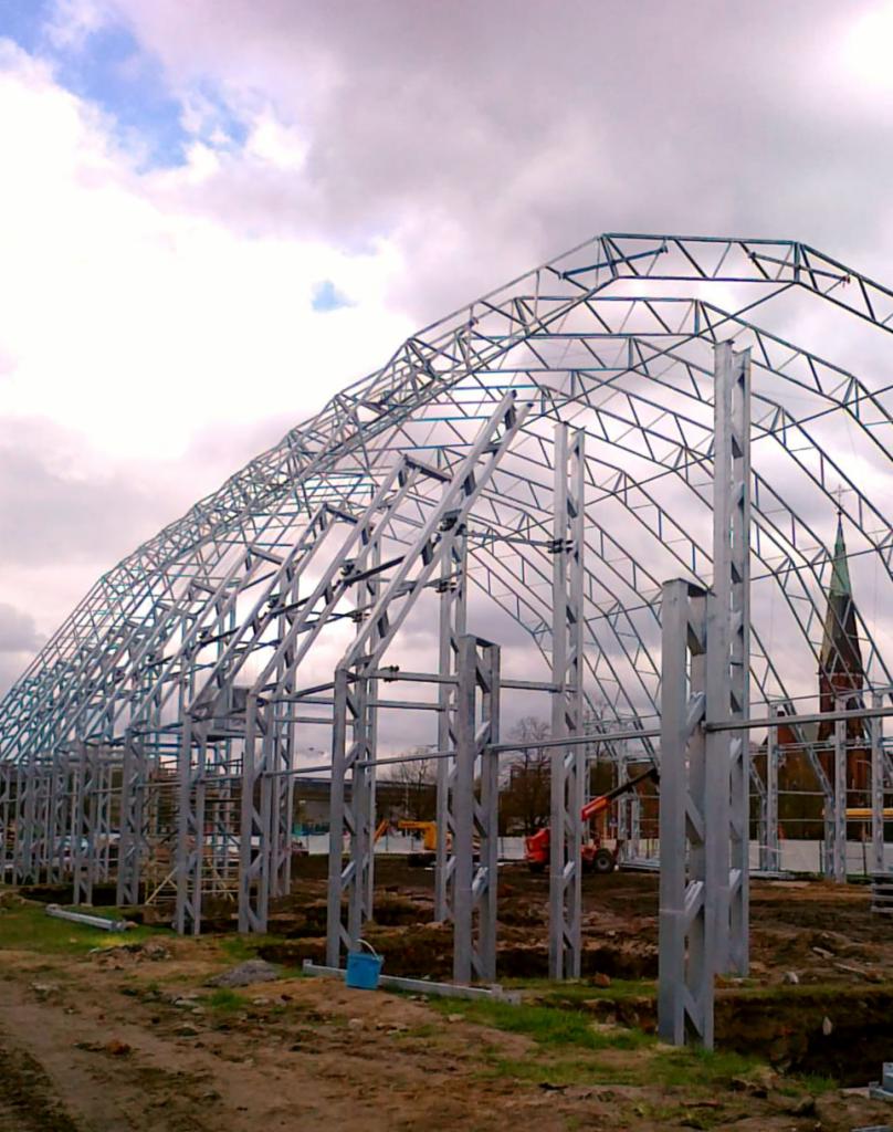Wznoszenie szkieletu hali: widok na stalową konstrukcję z pionowymi słupami i łukowym dachem w trakcie montażu, z widocznym sprzętem budowlanym w tle i fragmentem kościoła w oddali.