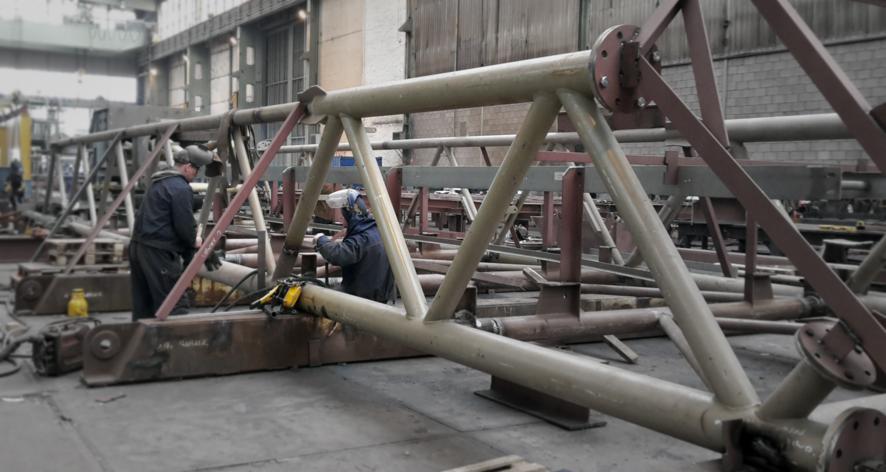 Spawanie elementu kratownicy z rur stalowych w hali produkcyjnej, widoczny spawacz w kombinezonie i przyłbicy, elementy konstrukcji w kolorze beżowym i bordowym.