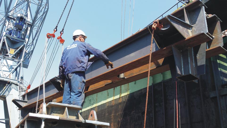 Pracownik w kasku i roboczym ubraniu ZUSO, stojący na platformie, nadzoruje montaż stalowej belki konstrukcyjnej przy użyciu żurawia na tle błękitnego nieba.