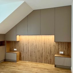 FHU JACK - Zabudowa wnęki na poddaszu z drewnianym panelem i szafkami z oświetleniem LED. Widoczne gniazdka elektryczne i drewniana podłoga. Realizacja pod wymiar.