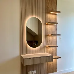 Nowoczesna toaletka z owalnym lustrem podświetlanym LED i drewnianymi półkami z oświetleniem punktowym, zamontowana na ścianie w jasnym wnętrzu.