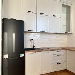 FHU JACK - Biała kuchnia z czarnym zlewem i lodówką Candy. Klasyczne frezowane fronty, drewniany blat i podłoga. Górne szafki częściowo przeszklone.