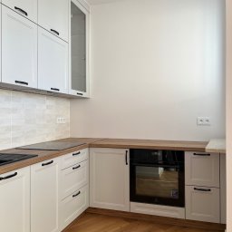 FHU JACK - Biała kuchnia w stylu klasycznym z drewnianym blatem i podłogą. Widoczny piekarnik, płyta grzewcza i szafki z czarnymi uchwytami. Jasne płytki na ścianie.