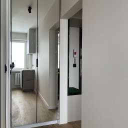 FHU JACK - Nowoczesna szafa wnękowa z lustrzanymi drzwiami, minimalistyczne uchwyty, widok na jasną kuchnię w tle, drewniana podłoga, białe ściany.