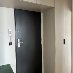 FHU JACK - Nowoczesne wykończenie przedpokoju: czarne drzwi wejściowe, zabudowa szafy w odcieniach szarości, zielone siedzisko. Minimalistyczny design, jasne panele podłogowe.