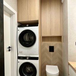 FHU JACK Jacek Zabłocki - Kompaktowa łazienka z zabudową z jasnego drewna, integrującą pralkę Samsung i suszarkę Hotpoint w słupku. Obok podwieszana toaleta na tle beżowych płytek w jodełkę.
