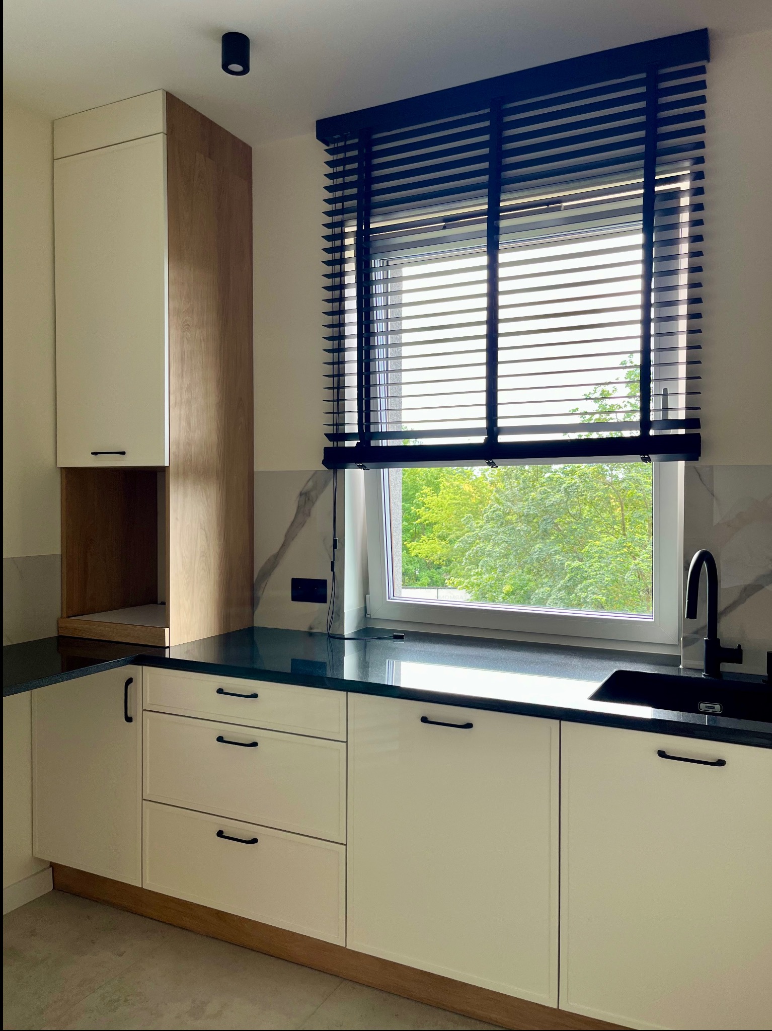 Nowoczesna kuchnia z białymi frontami, czarnym blatem i armaturą. Okno z drewnianymi żaluzjami. Minimalistyczny design i naturalne światło.