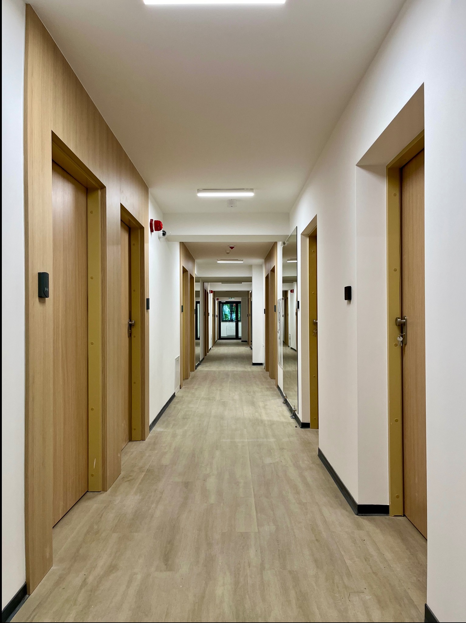 Długi korytarz w budynku publicznym z drewnianymi drzwiami i panelami ściennymi, wykończony w jasnych barwach, z widocznymi elementami bezpieczeństwa pożarowego.