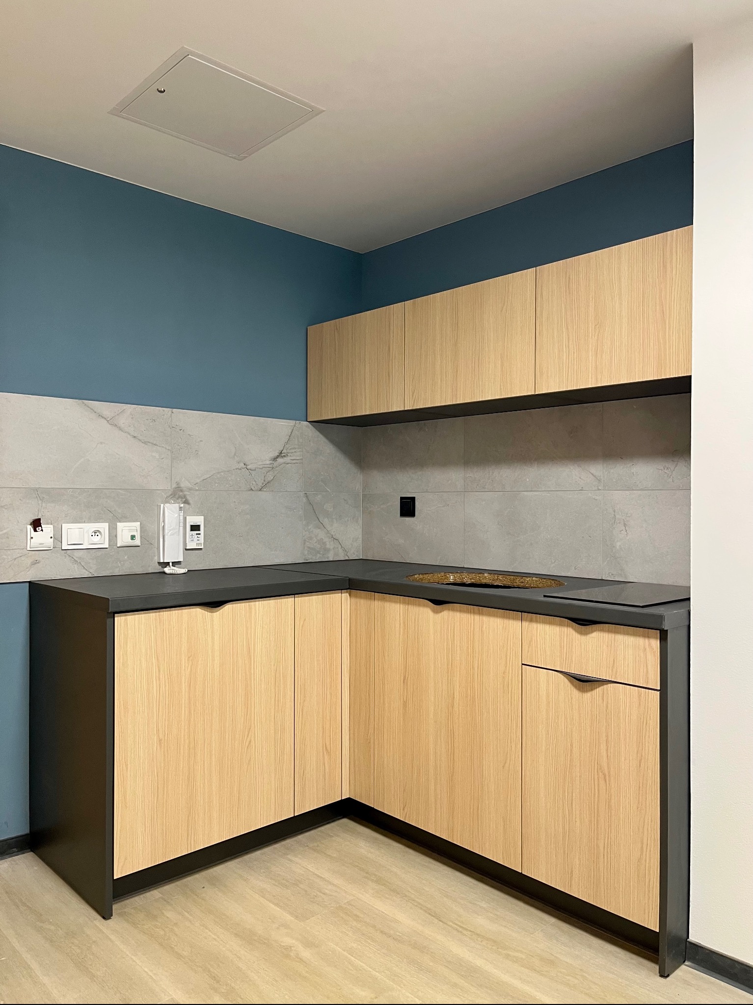 Jasna, drewniana zabudowa kuchenna w aneksie z szarym blatem i płytkami. Niebieska ściana, widoczne gniazdka i domofon. Prosty, nowoczesny design.