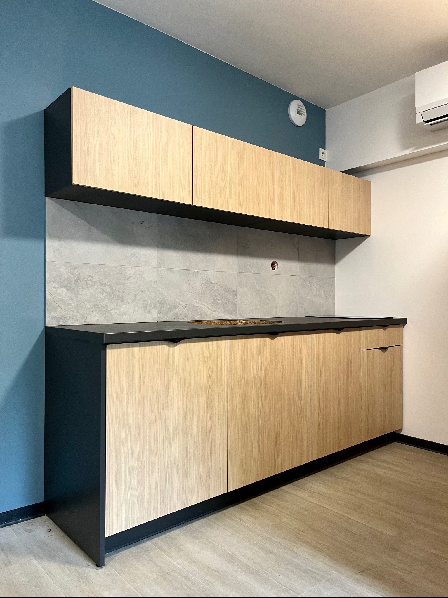 Jasna, nowoczesna kuchnia z drewnianymi frontami i czarnym blatem. Błękitna ściana, szare płytki nad blatem. Widoczny fragment podłogi i klimatyzacja.