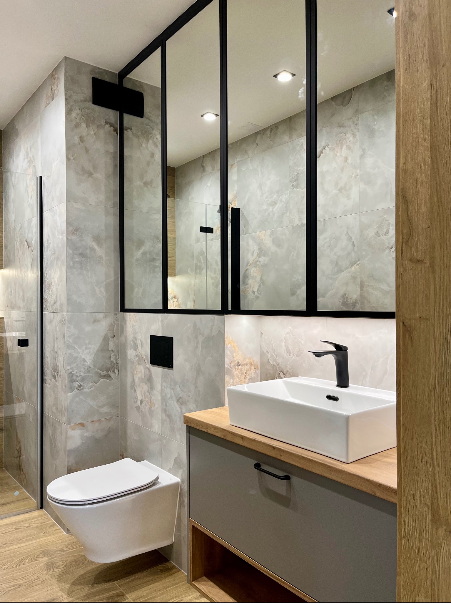 Nowoczesna łazienka z szarymi płytkami imitującymi marmur, wiszącą toaletą i umywalką nablatową na szafce z blatem drewnianym. Kabina prysznicowa ze szklanymi drzwiami.