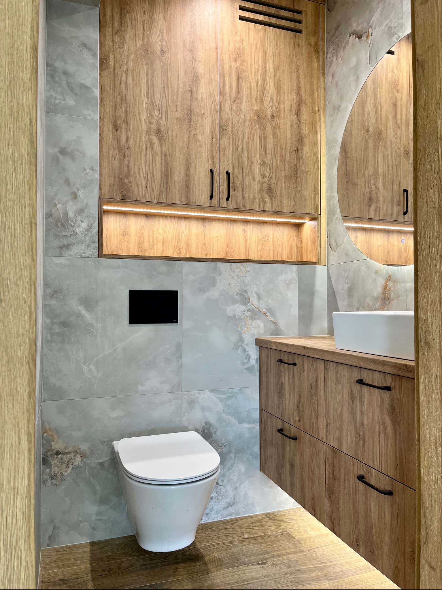 Nowoczesna łazienka z drewnianymi szafkami, okrągłym lustrem z podświetleniem LED i marmurowymi płytkami. Wisząca toaleta i drewnopodobna podłoga.