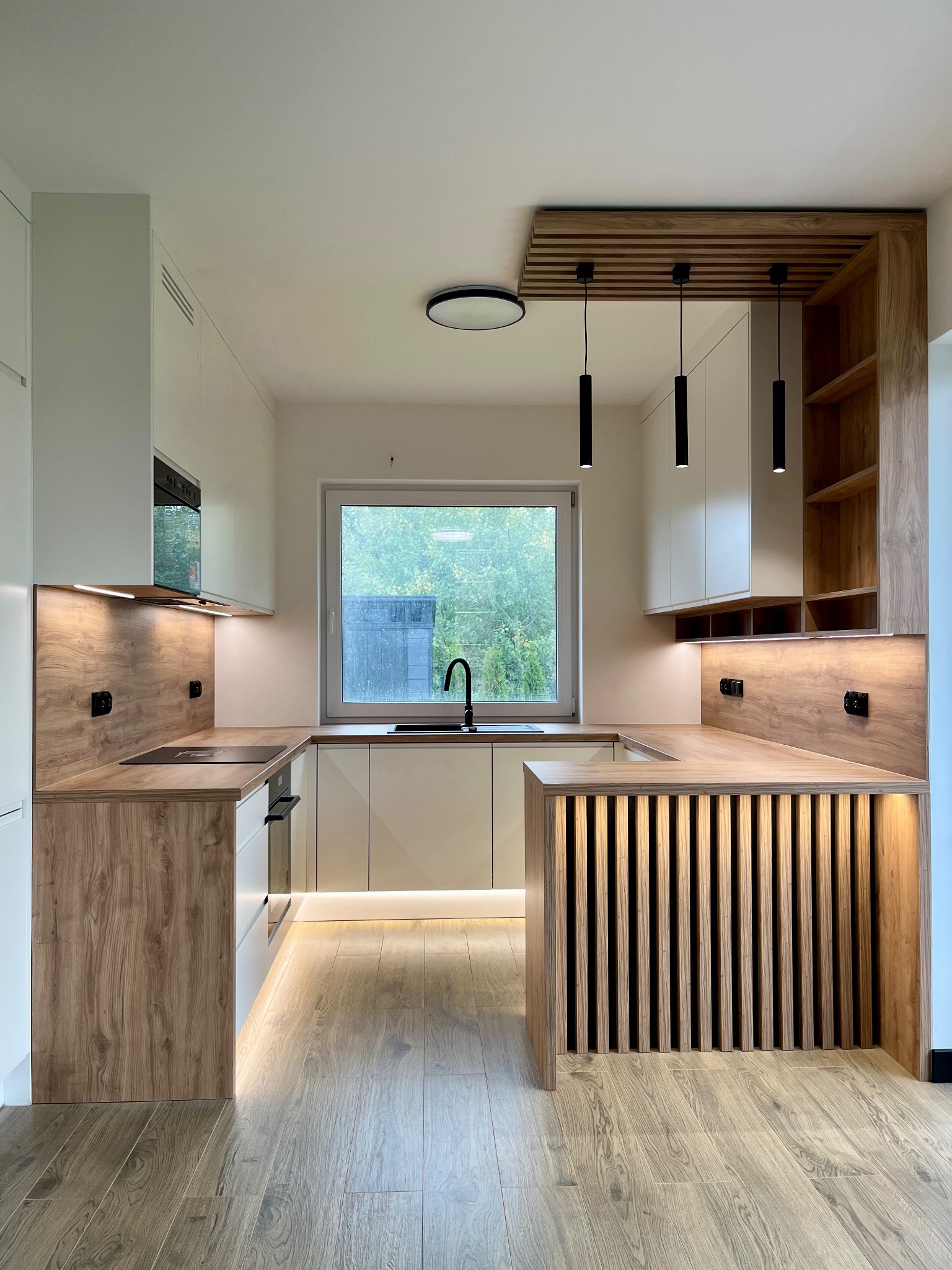 Nowoczesna kuchnia z drewnianymi akcentami: wyspa z lamelami, oświetlenie LED pod szafkami, czarne lampy wiszące i armatura, okno z widokiem na zieleń.