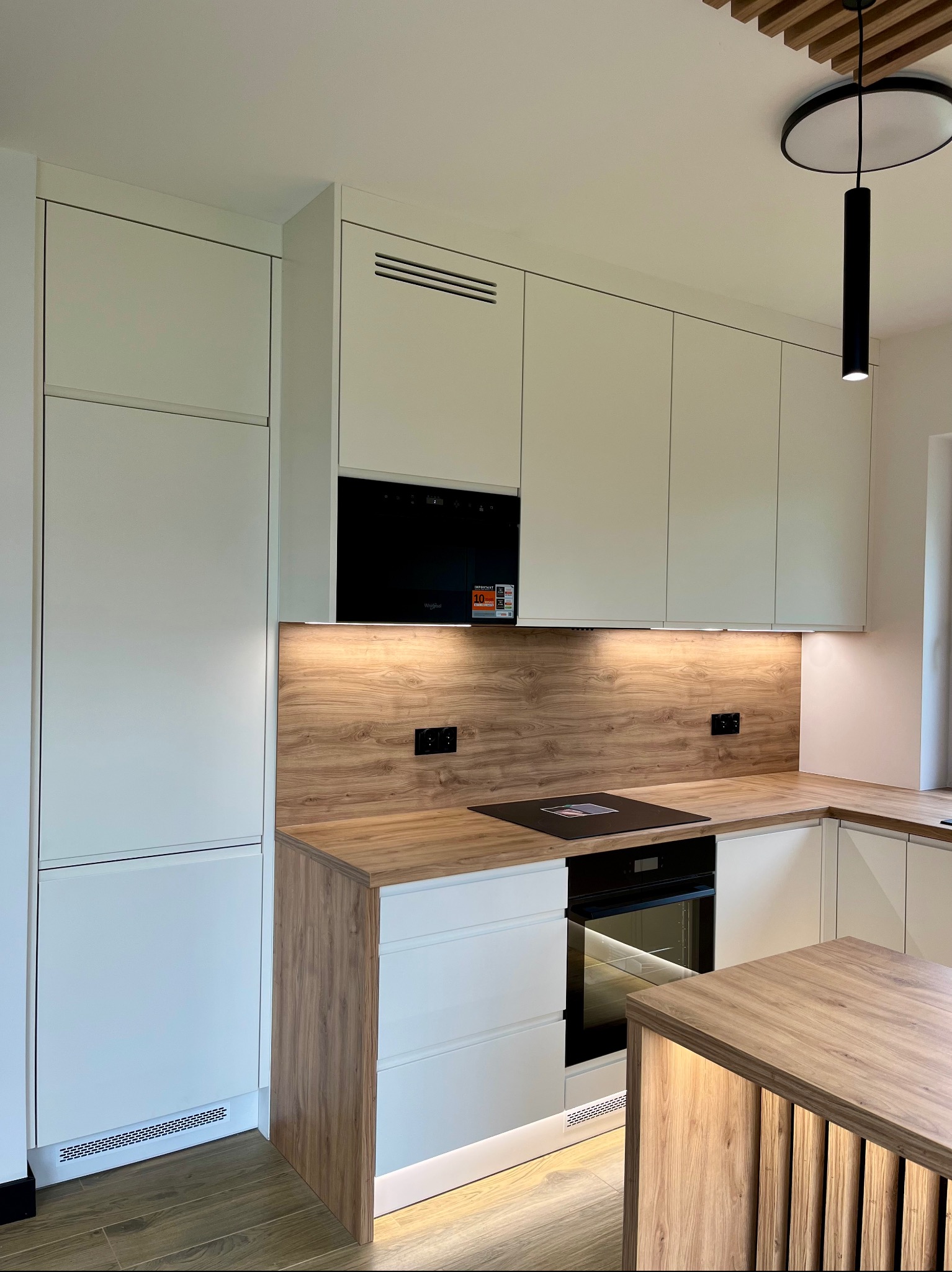 Nowoczesna kuchnia z białymi szafkami i drewnianym blatem. Na ścianie płyta imitująca drewno, oświetlenie podszafkowe. Widoczny piekarnik i płyta indukcyjna.