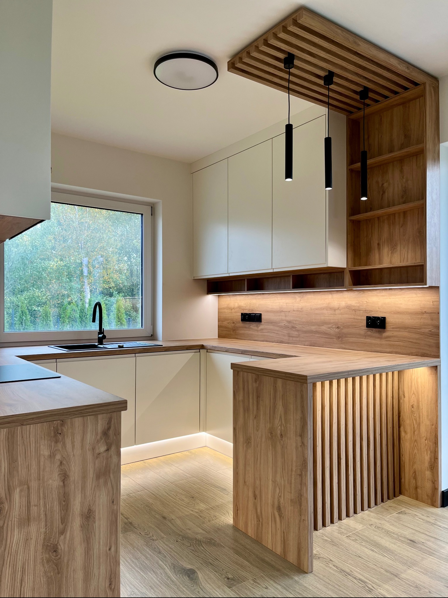 Nowoczesna kuchnia z drewnianymi akcentami: wyspa z lamelami, beżowe szafki, czarne lampy wiszące i okno z widokiem na zieleń. Oświetlenie LED pod szafkami.