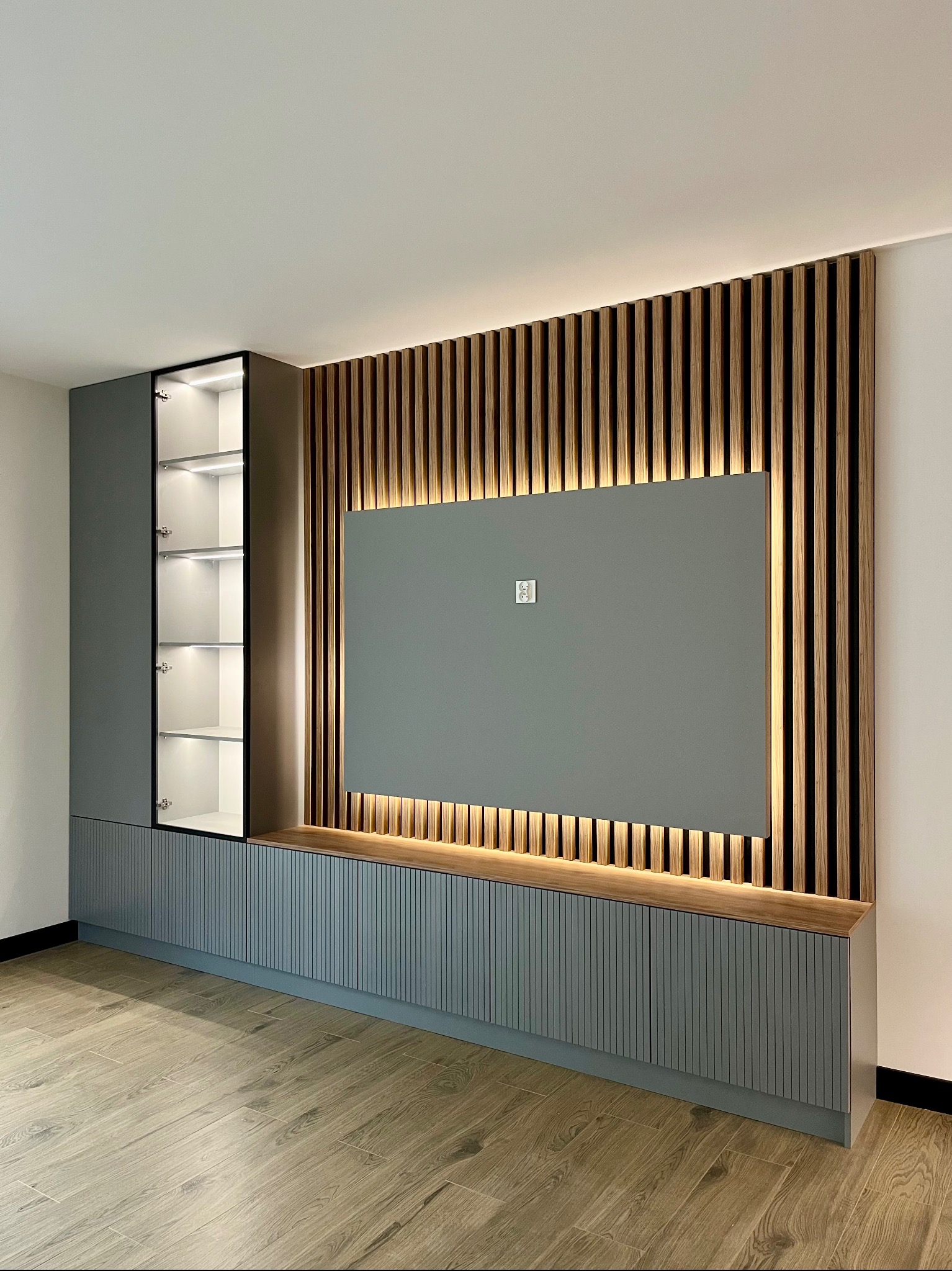 Szara zabudowa meblowa z oświetleniem LED i drewnianymi lamelami na ścianie. Otwarta szafka z szklanymi półkami. Nowoczesny design, jasna podłoga.