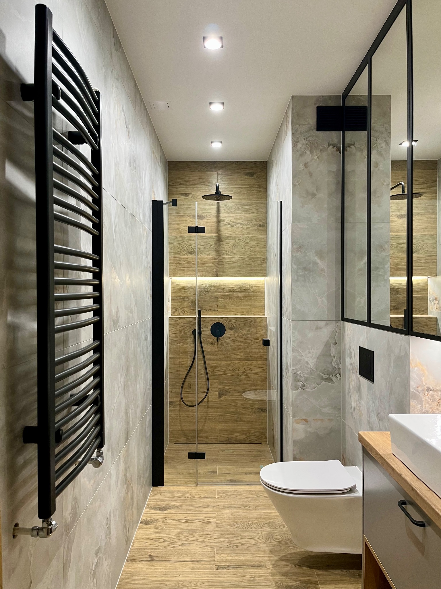 Nowoczesna łazienka z kabiną prysznicową typu walk-in, grzejnikiem drabinkowym i umywalką z szafką. Uwagę zwracają płytki imitujące marmur i drewno.