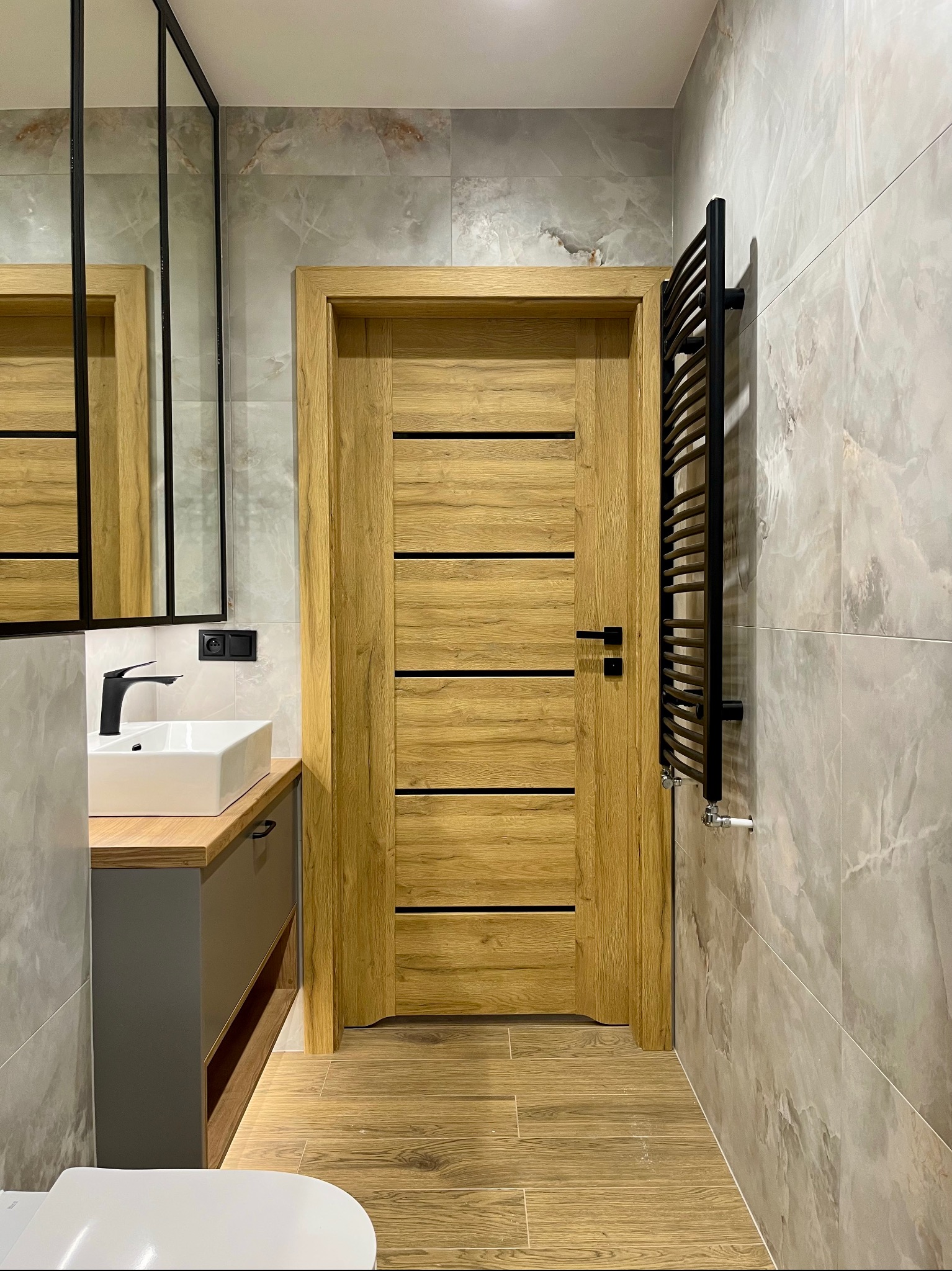 Nowoczesna łazienka z drewnianymi akcentami: drzwi z poziomymi panelami, szafka pod umywalką i grzejnik drabinkowy na tle marmurowych płytek.