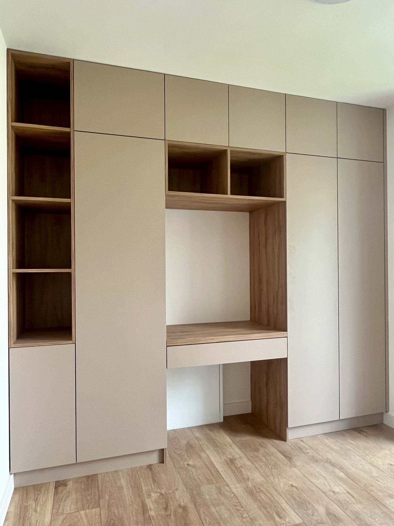 Beżowa szafa wnękowa z drewnianym biurkiem i otwartymi półkami, dopasowana do wnętrza. Nowoczesny design, jasne drewno na podłodze.