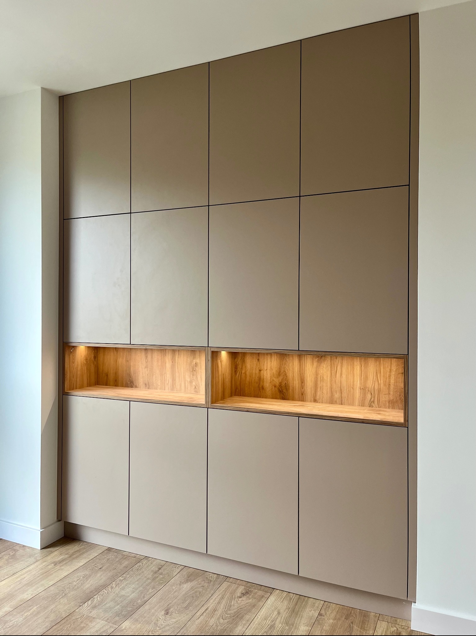 Beżowa szafa wnękowa z drewnianymi, podświetlonymi wnękami, dopasowana do jasnego wnętrza z drewnianą podłogą. Minimalistyczny design.