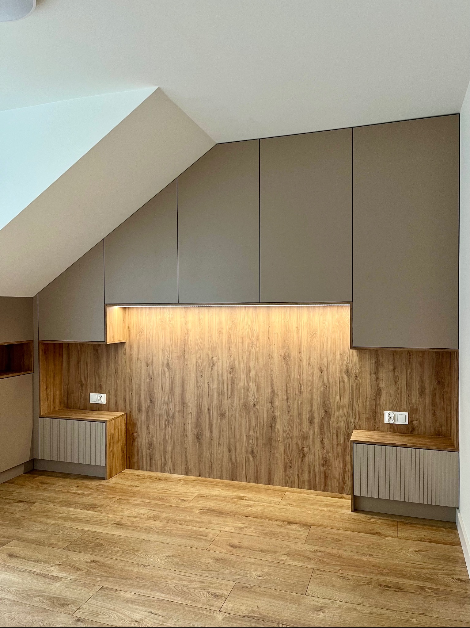 Zabudowa wnęki na poddaszu z drewnianym panelem i szafkami z oświetleniem LED. Widoczne gniazdka elektryczne i drewniana podłoga. Realizacja pod wymiar.