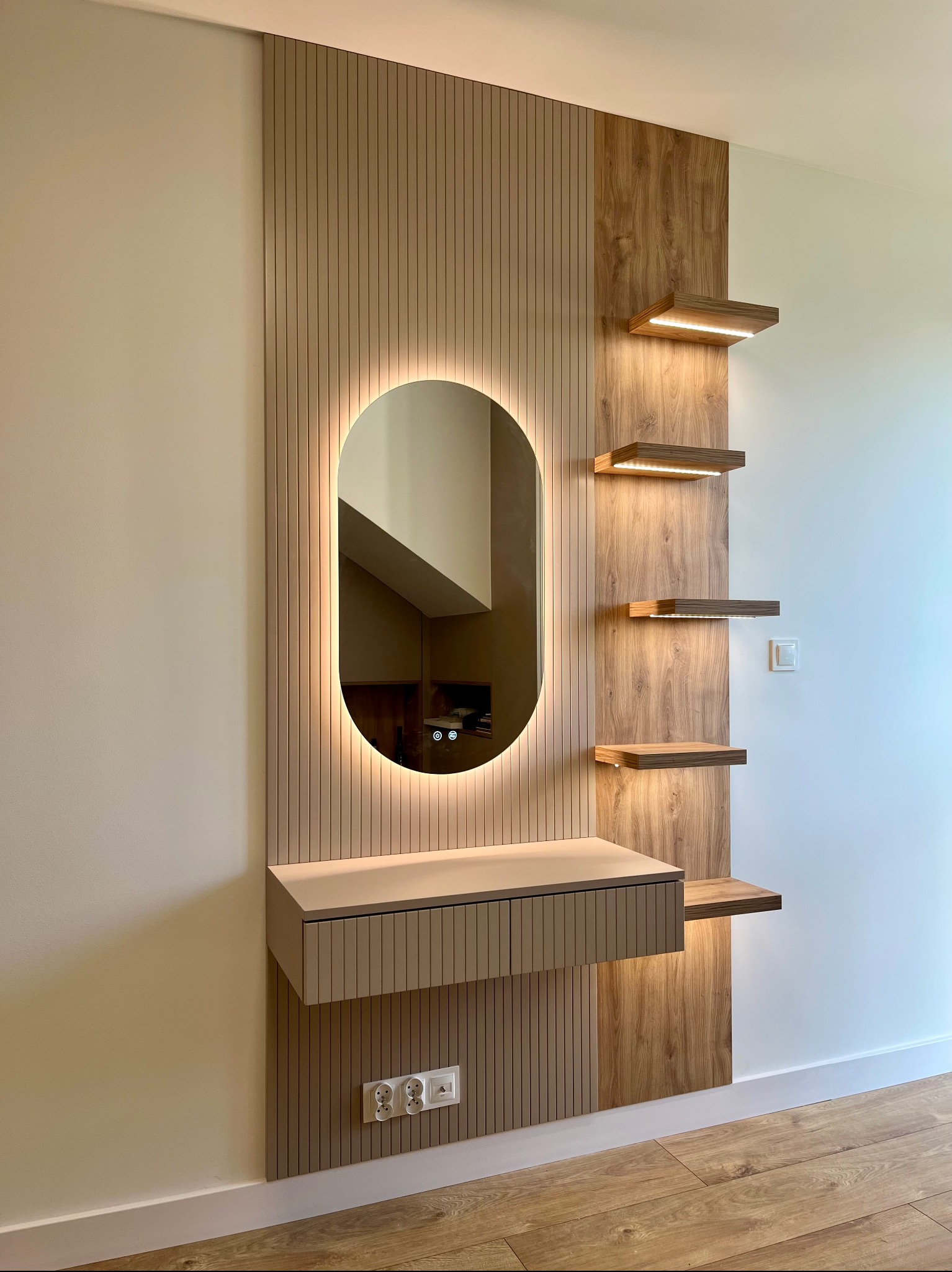 Nowoczesna toaletka z owalnym lustrem podświetlanym LED i drewnianymi półkami z oświetleniem punktowym, zamontowana na ścianie w jasnym wnętrzu.