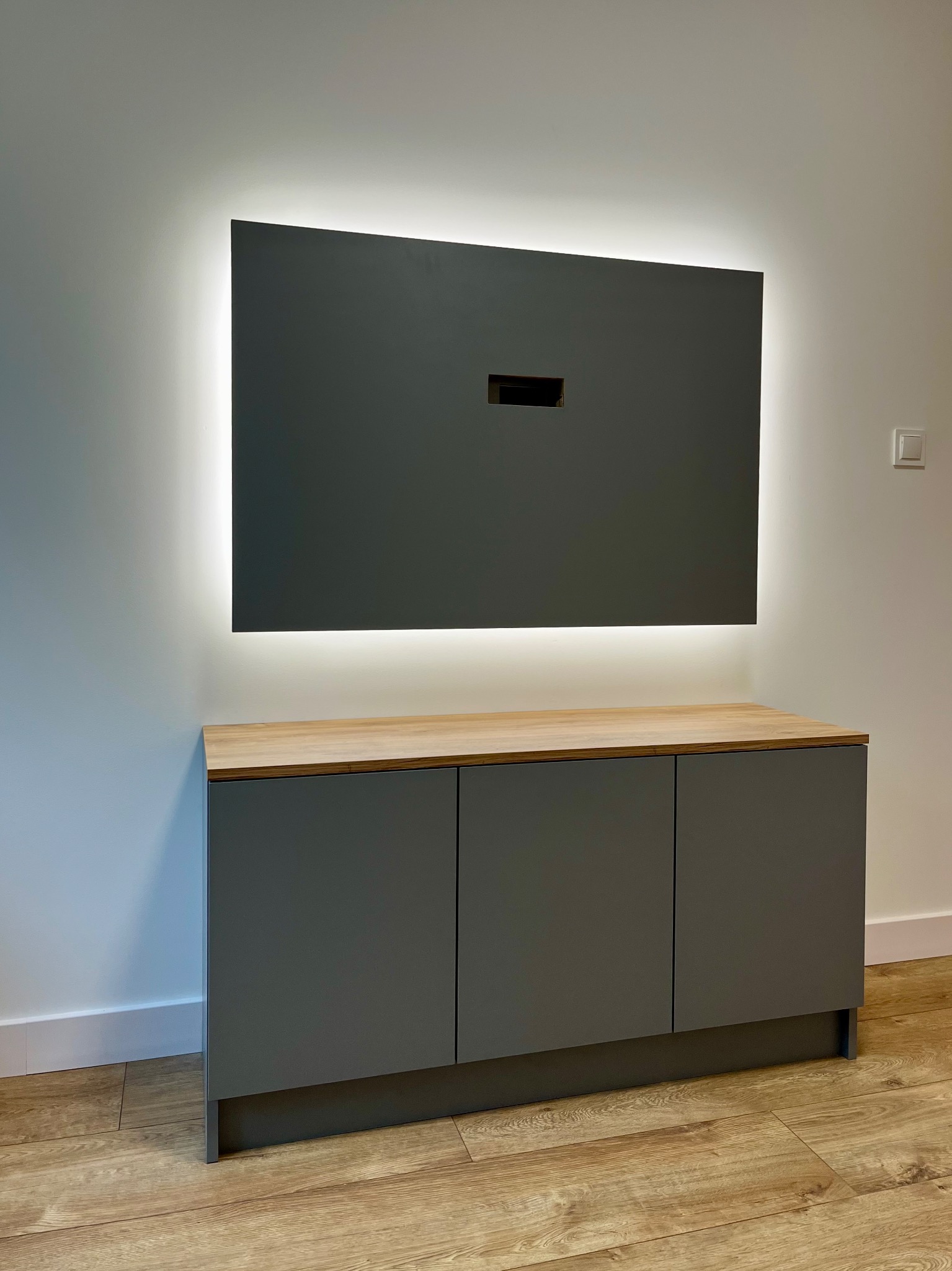 Nowoczesna szafka RTV w szarym kolorze z drewnianym blatem i podświetleniem LED, zamontowana na ścianie. Minimalistyczny design, jasne wnętrze.