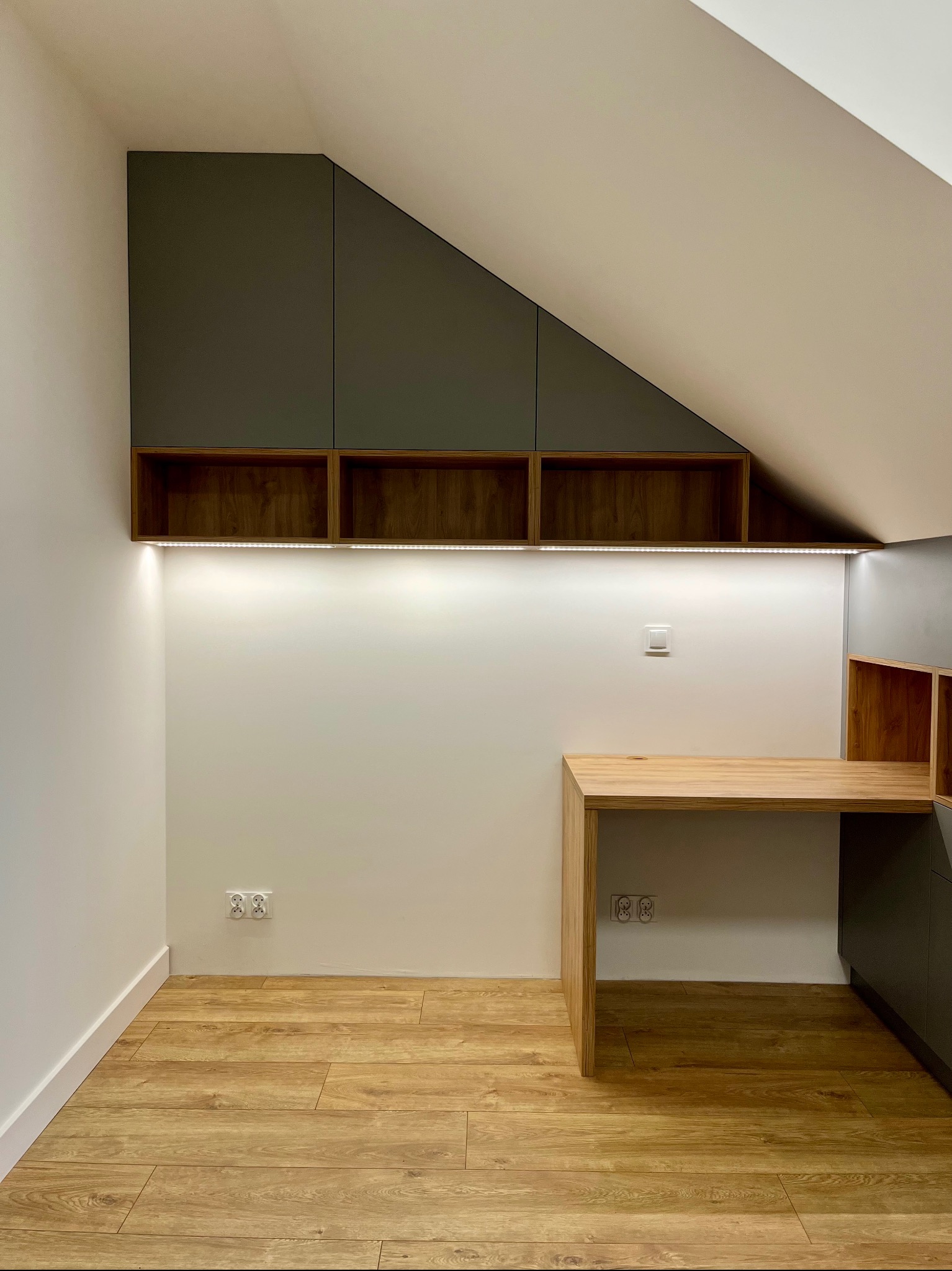 Funkcjonalne biuro w skosie poddasza z szafkami i blatem z drewna. Oświetlenie LED podkreśla unikalny charakter wnętrza. Drewniana podłoga dodaje ciepła.