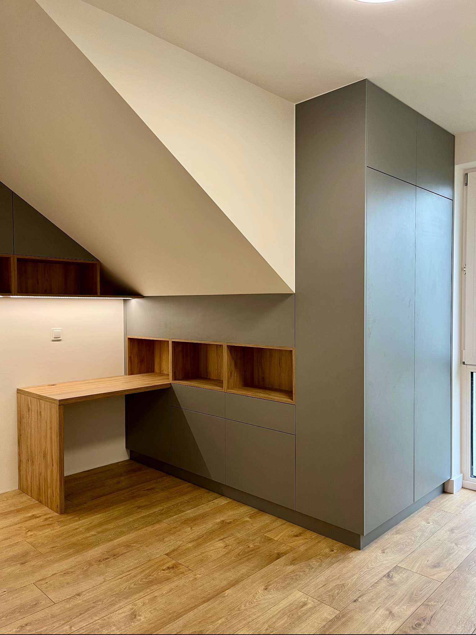 Funkcjonalna zabudowa pod skosem: szara szafa, drewniane półki i biurko z oświetleniem LED, dopasowane do podłogi. Estetyczne wykończenie wnętrza.