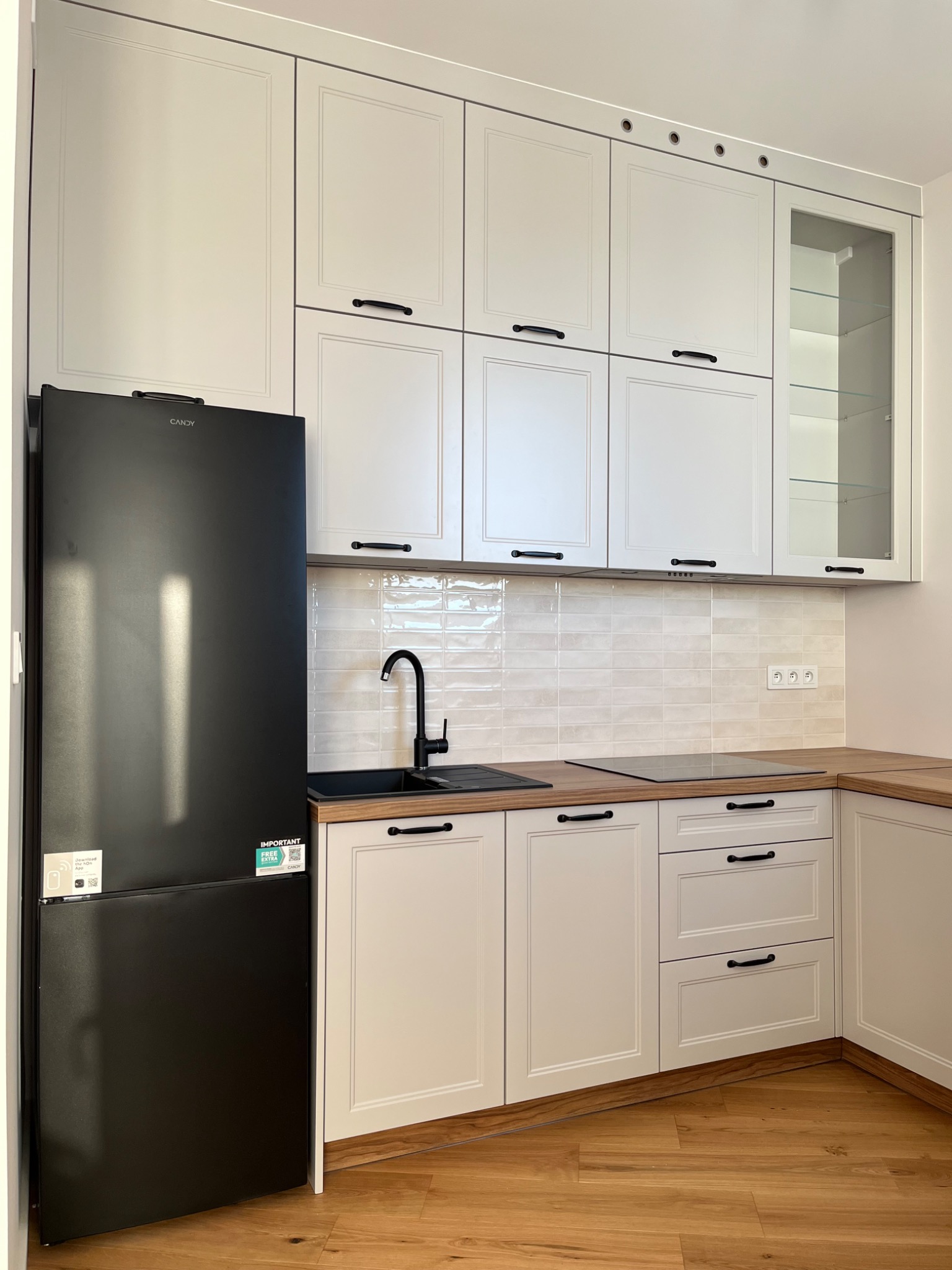 Biała kuchnia z czarnym zlewem i lodówką Candy. Klasyczne frezowane fronty, drewniany blat i podłoga. Górne szafki częściowo przeszklone.