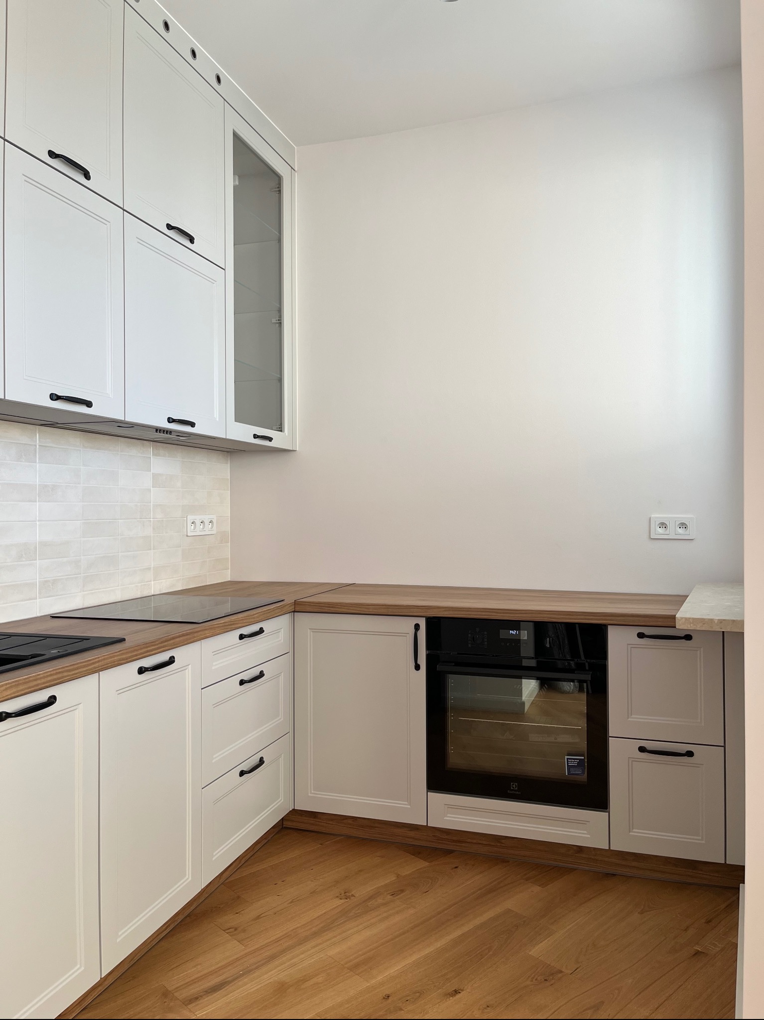 Biała kuchnia w stylu klasycznym z drewnianym blatem i podłogą. Widoczny piekarnik, płyta grzewcza i szafki z czarnymi uchwytami. Jasne płytki na ścianie.