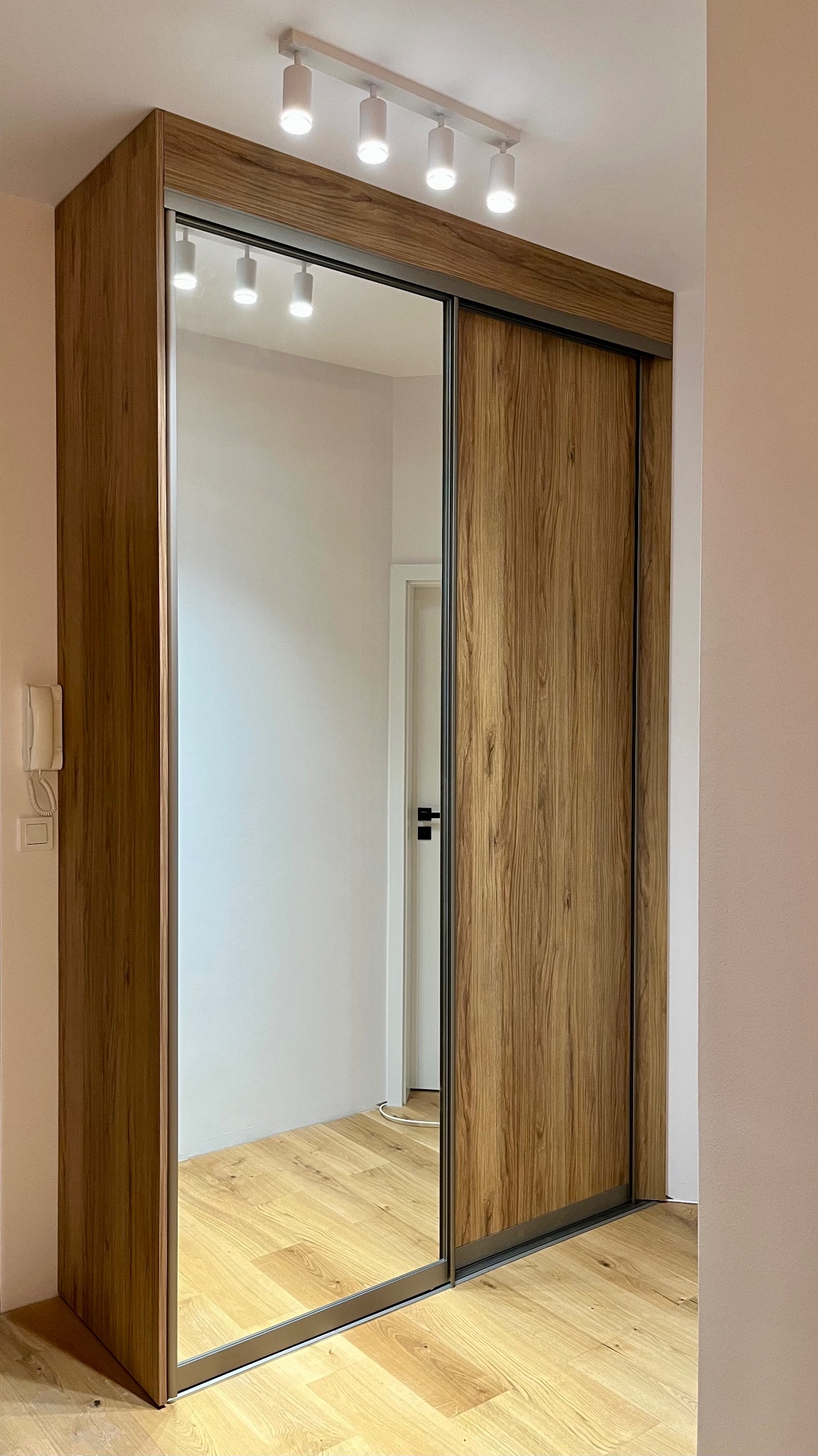 Szafa wnękowa z przesuwnymi drzwiami lustrzanymi i drewnianym wykończeniem, oświetlona listwą LED, na tle jasnego wnętrza z parkietem.