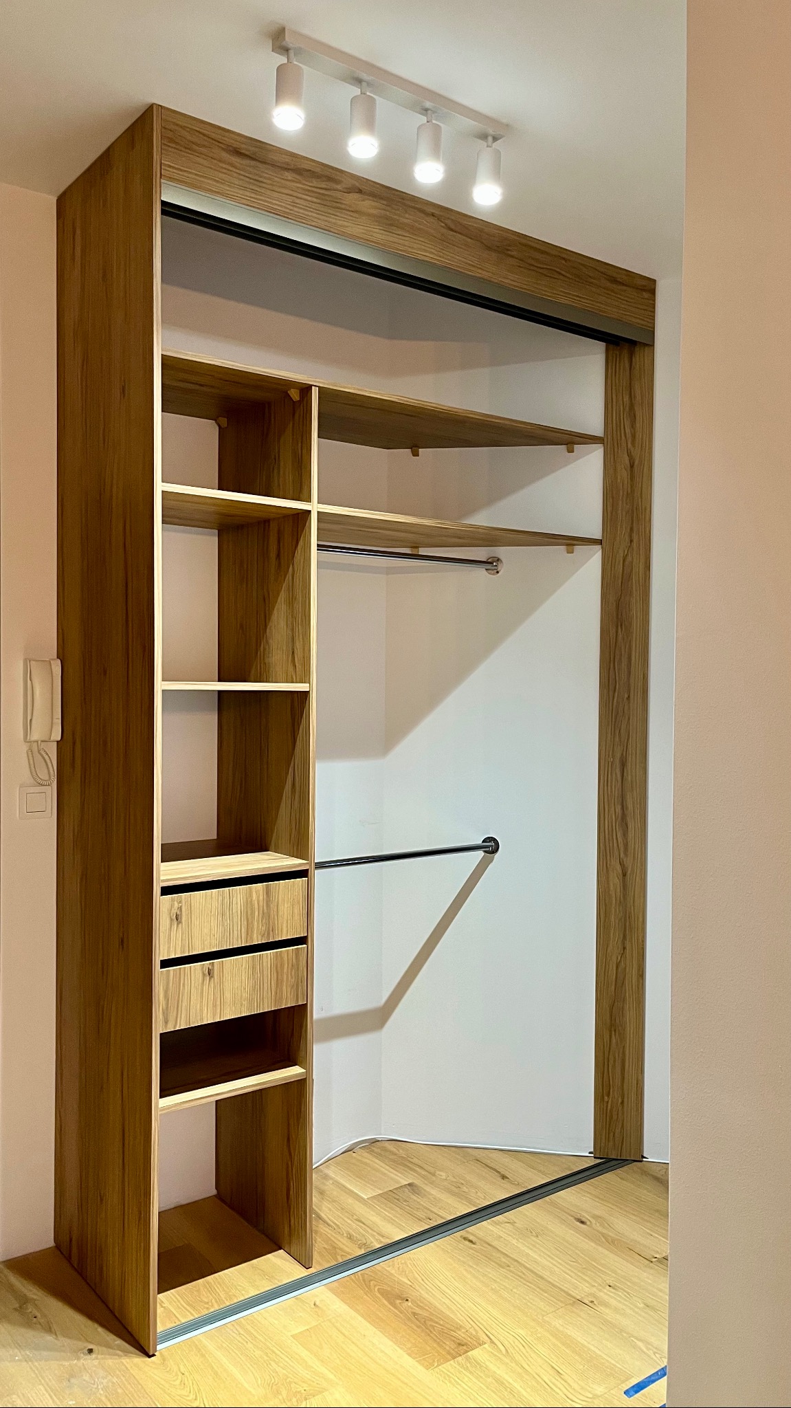 Drewniana szafa wnękowa z przesuwnymi drzwiami, oświetleniem punktowym i półkami. Widoczna podłoga z paneli. Ujęcie z perspektywy wnętrza szafy.