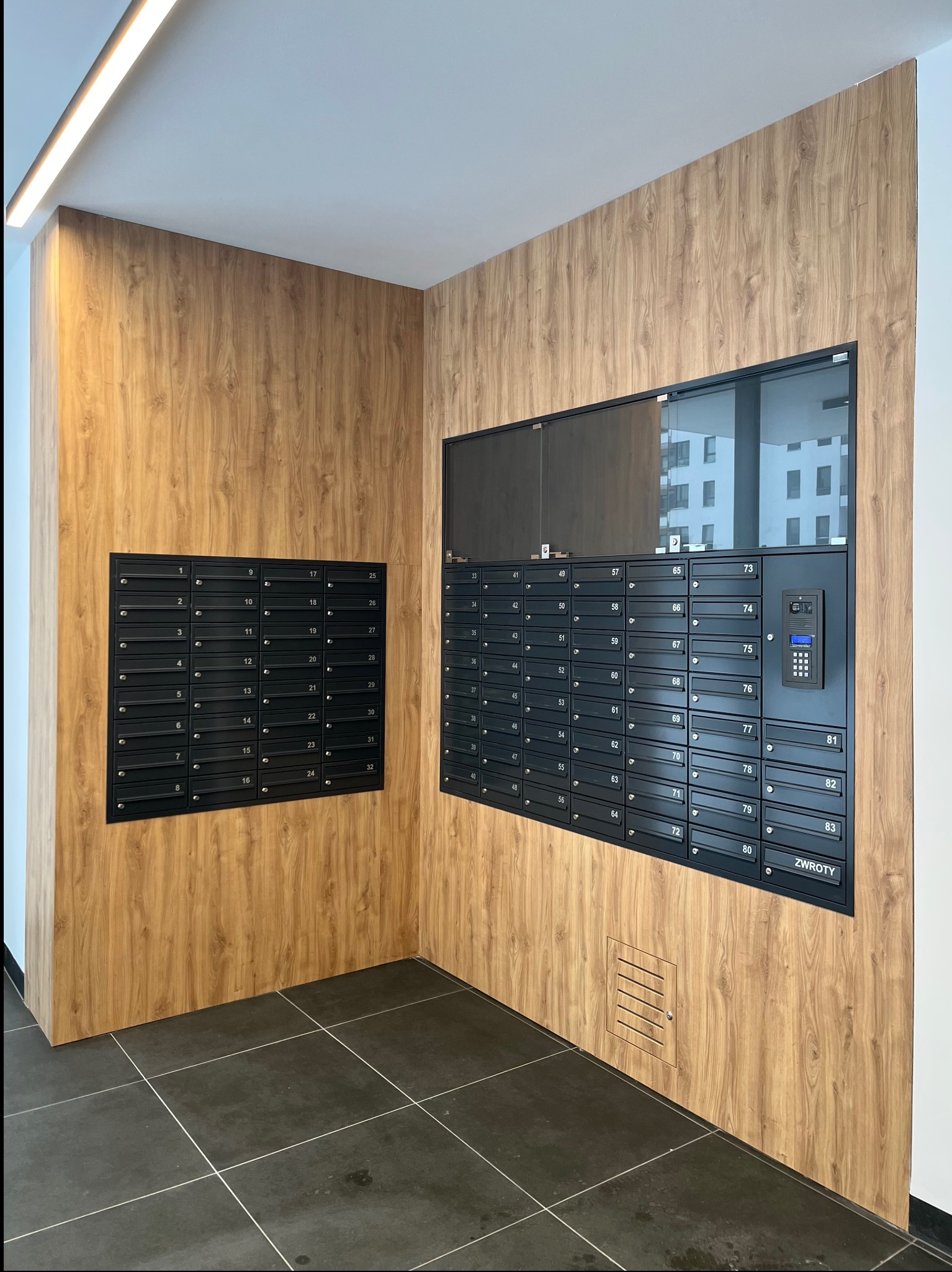Wnętrze klatki schodowej z panelami drewnopodobnymi i czarnymi skrzynkami pocztowymi z numerami. Widoczny domofon i szara podłoga z płytek.