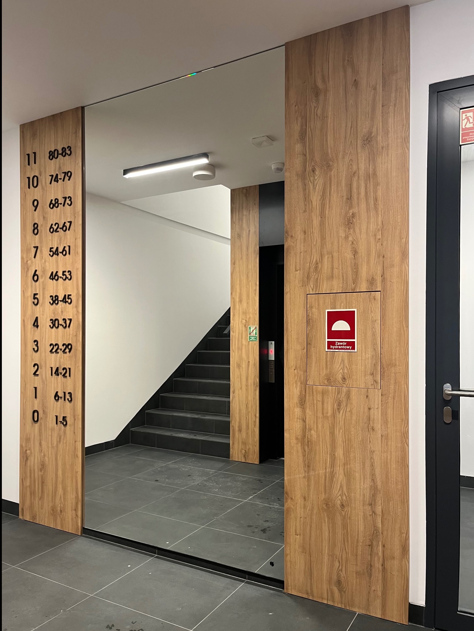 Wykończenie klatki schodowej w Olsztynie: drewnopodobne panele ścienne z numeracją mieszkań, szara podłoga, czarne schody i drzwi. Nowoczesny design.
