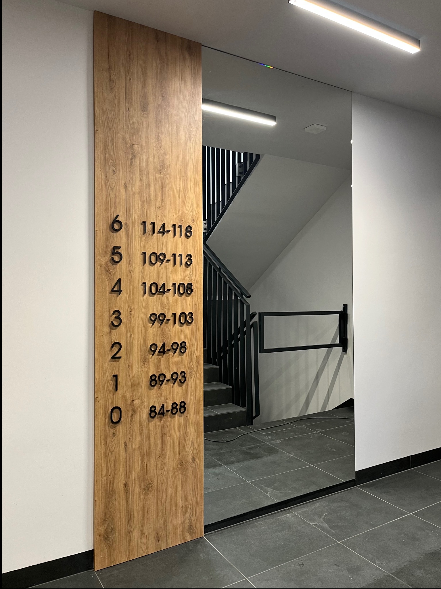 Tablica z numeracją mieszkań na drewnianej płycie, umieszczona przy schodach w nowoczesnym budynku. Minimalistyczny design, czarne cyfry, szara podłoga.
