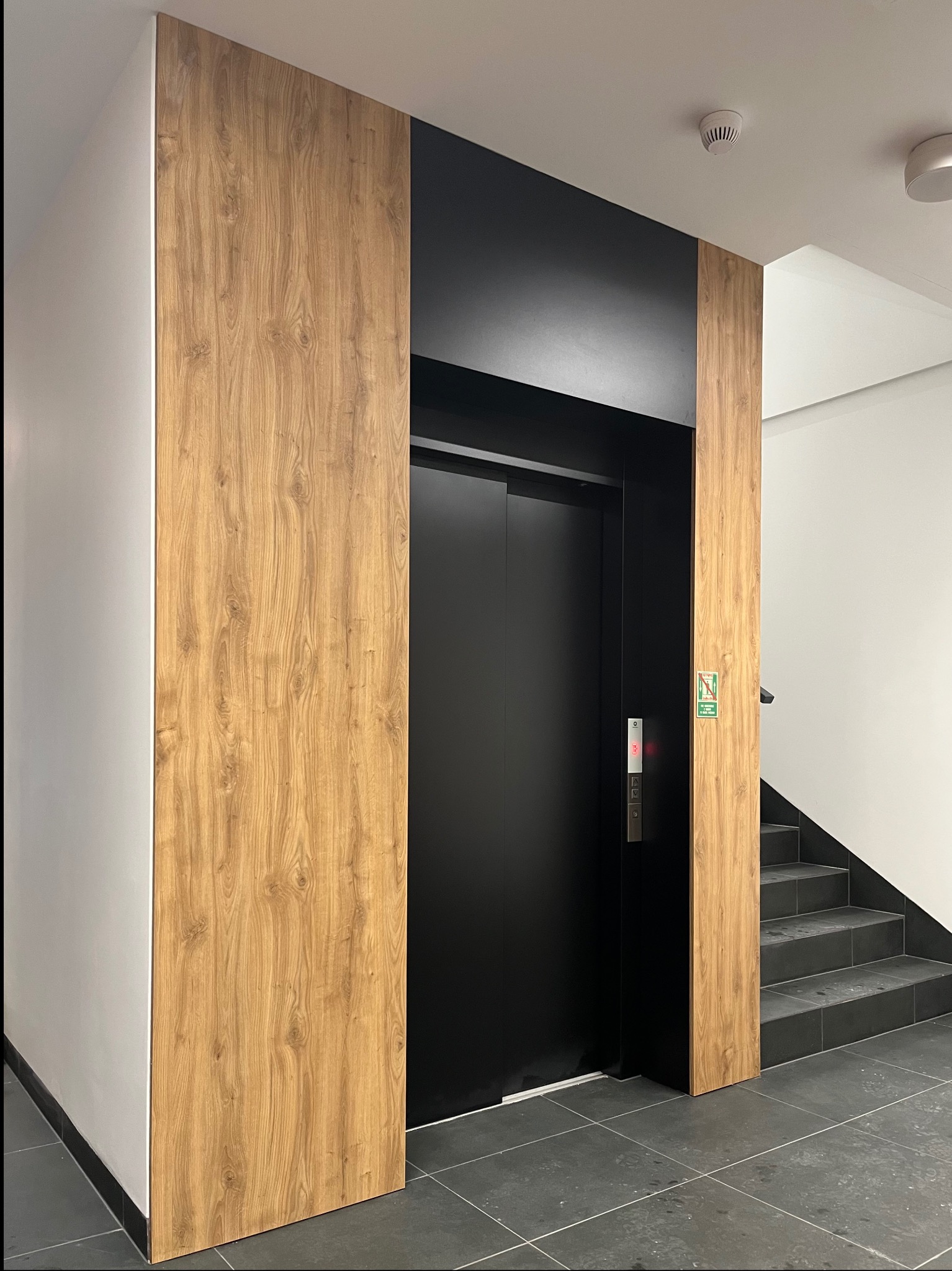 Nowoczesny hol z windą o czarnych drzwiach, obudowaną panelami z drewnopodobnym wzorem. Widoczne schody z ciemnymi płytkami i białe ściany.