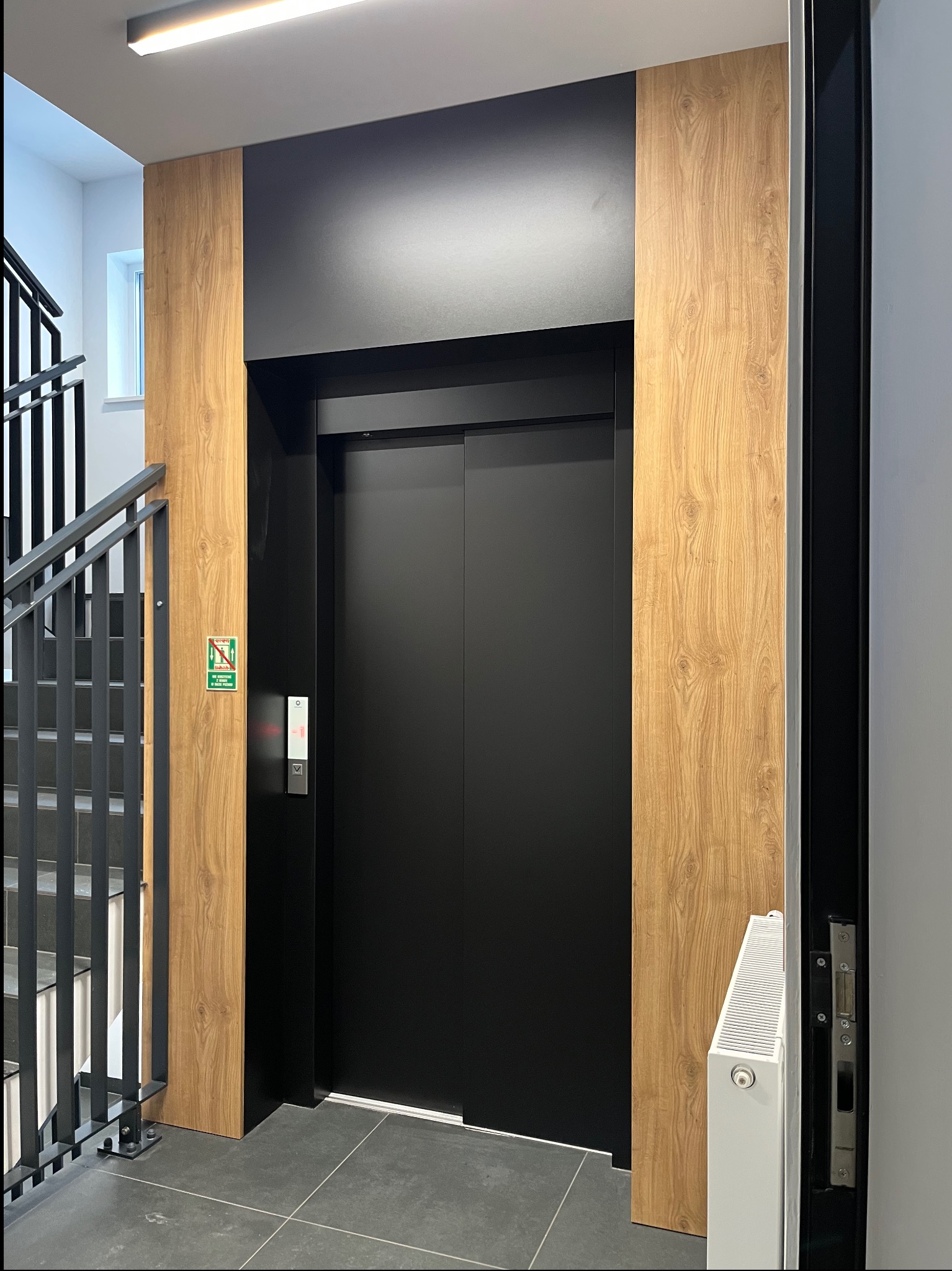 Nowoczesny, czarny dźwig osobowy z drewnianą obudową w klatce schodowej. Minimalistyczny design, szare płytki podłogowe, biały grzejnik.
