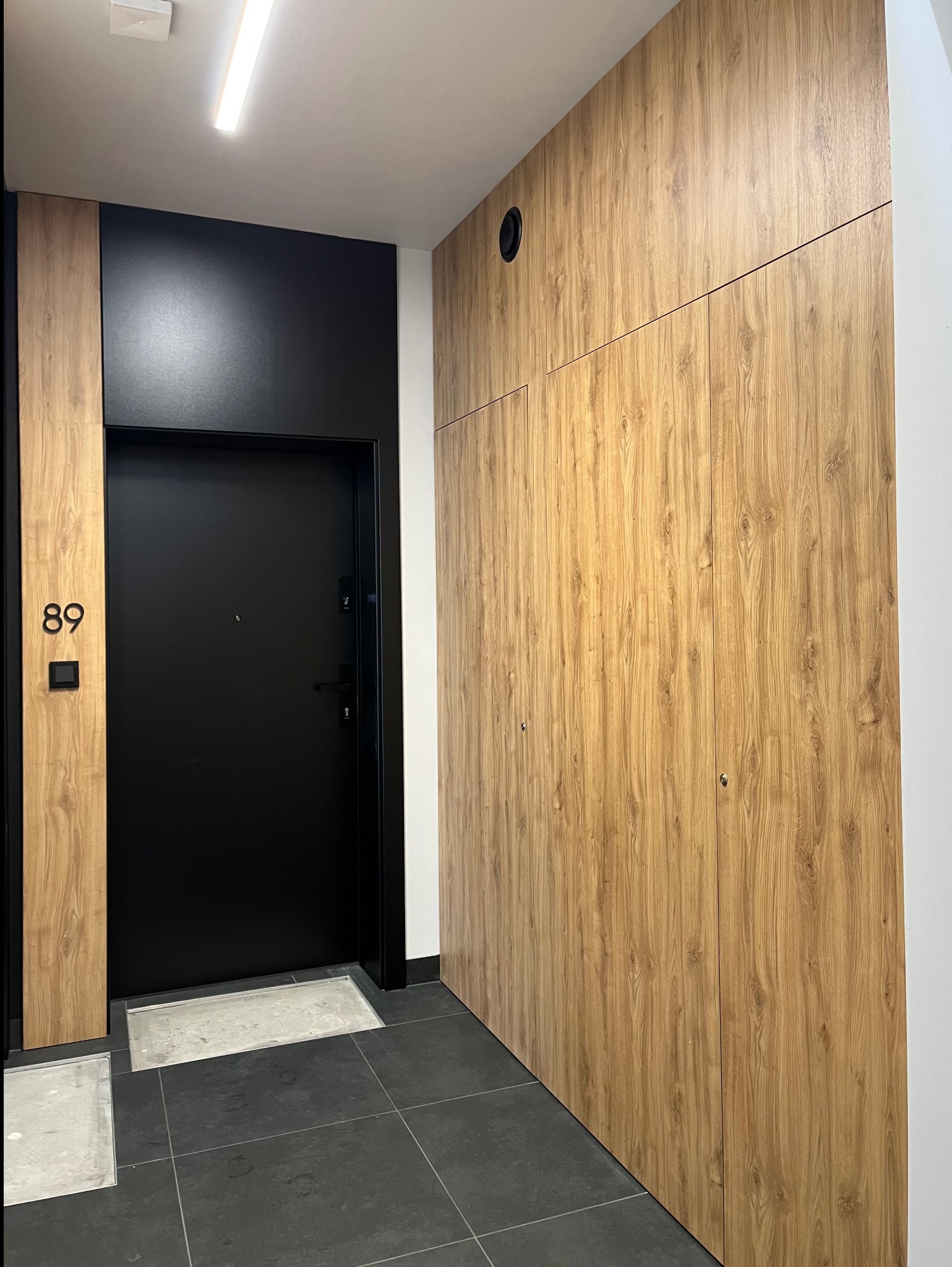 Nowoczesne wykończenie przedpokoju: czarne drzwi z numerem 89, drewnopodobna zabudowa szafy wnękowej od podłogi do sufitu, szara podłoga z płytek.