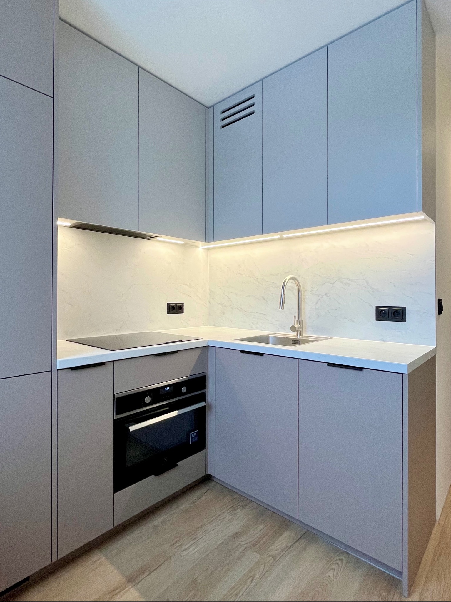 Nowoczesna kuchnia w odcieniach szarości z oświetleniem LED pod szafkami, marmurowym blatem i drewnianą podłogą. Minimalistyczny design z piekarnikiem i płytą indukcyjną.