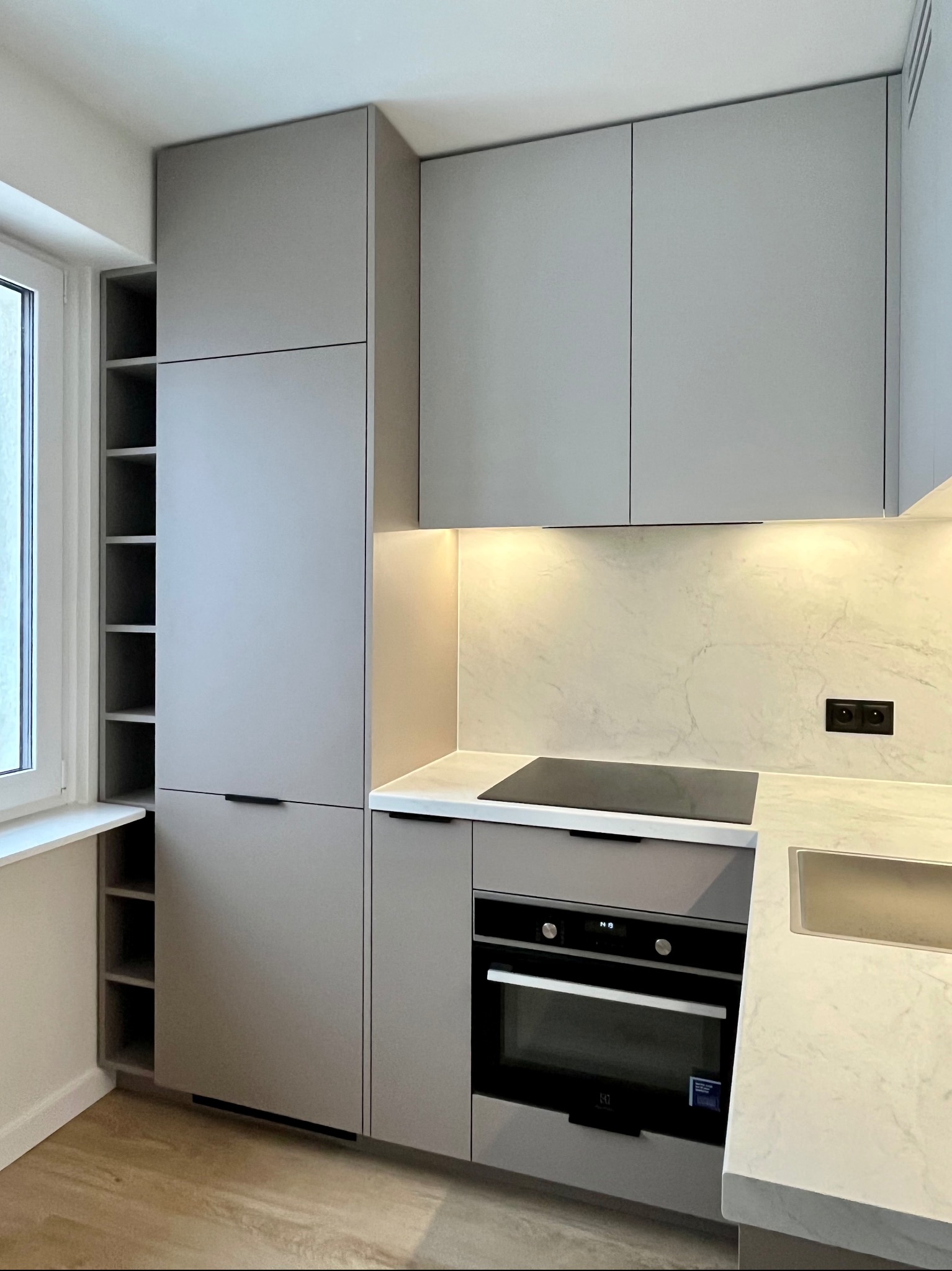 Nowoczesna kuchnia w odcieniach szarości z marmurowym blatem i oświetleniem LED. Widoczny piekarnik, płyta indukcyjna i regał wnękowy. Minimalistyczny design.