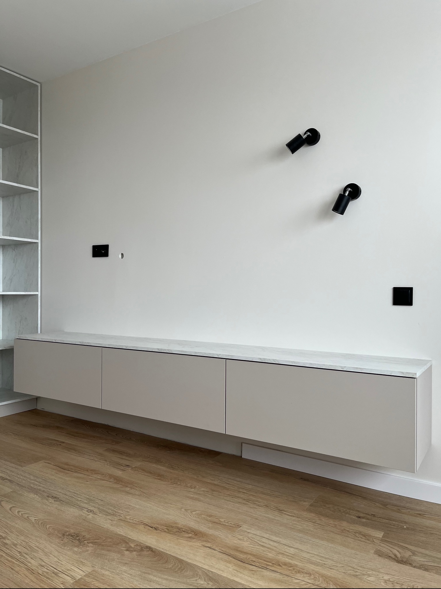 Minimalistyczny salon: beżowa szafka RTV z marmurowym blatem, czarne lampy punktowe na białej ścianie, drewniana podłoga. Nowoczesne wykończenie wnętrza.