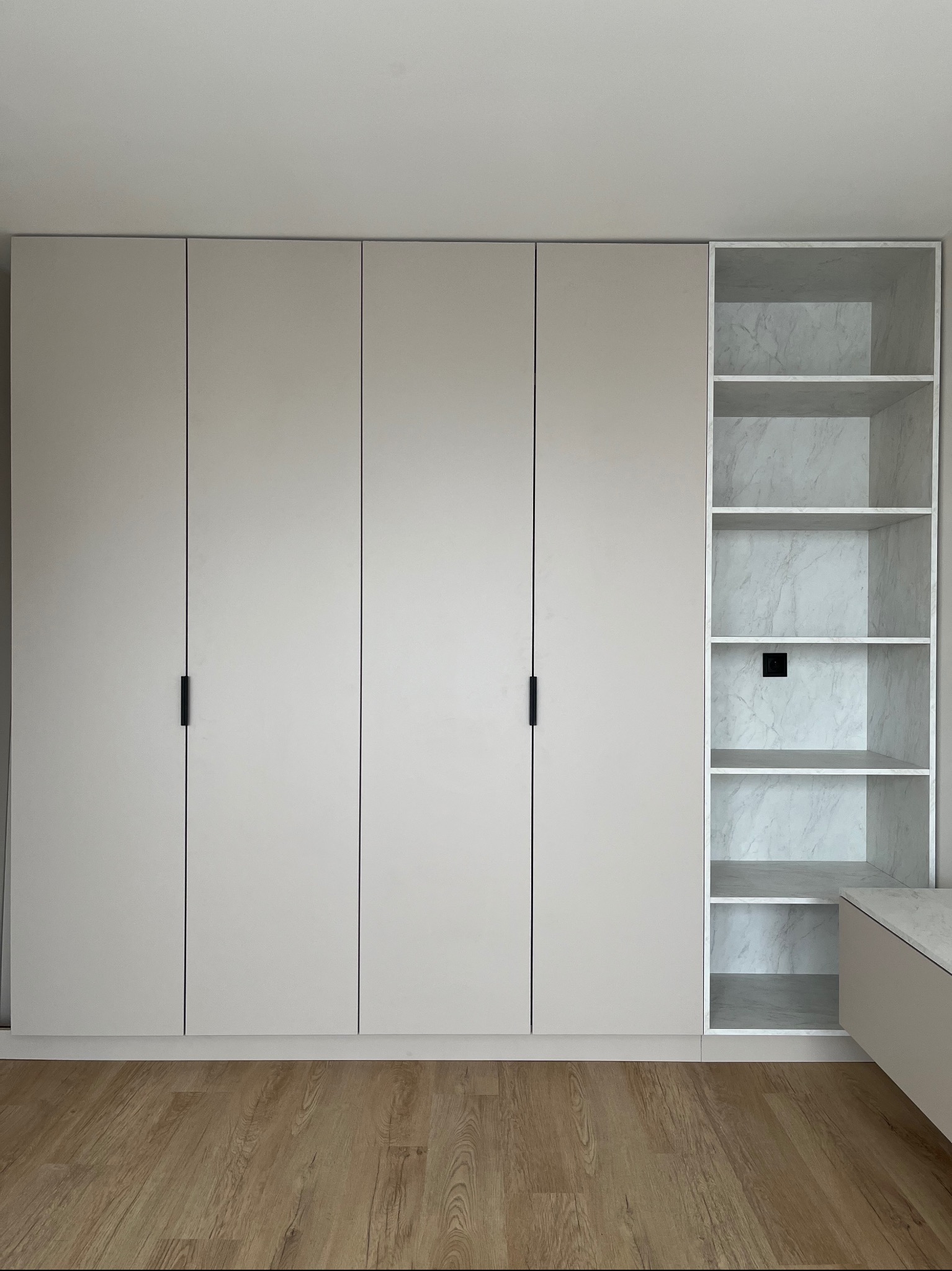 Jasna szafa wnękowa z czterema drzwiami i otwartą półką z marmurowym wzorem, minimalistyczne czarne uchwyty, drewniana podłoga, neutralne oświetlenie.