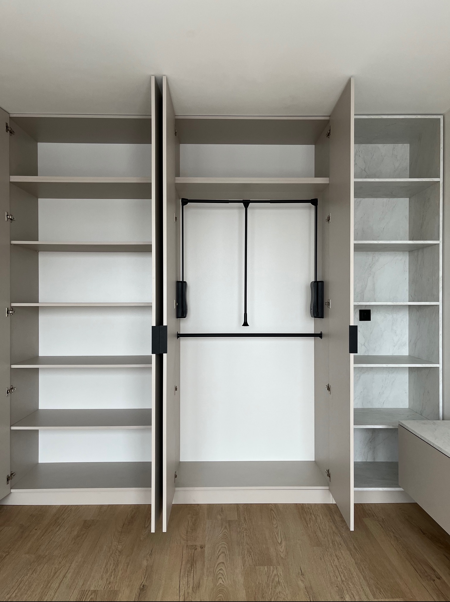 Otwarta, beżowa szafa wnękowa z półkami i drążkiem na ubrania, wykończona marmurowym wzorem na tylnej ścianie, na tle drewnianej podłogi.